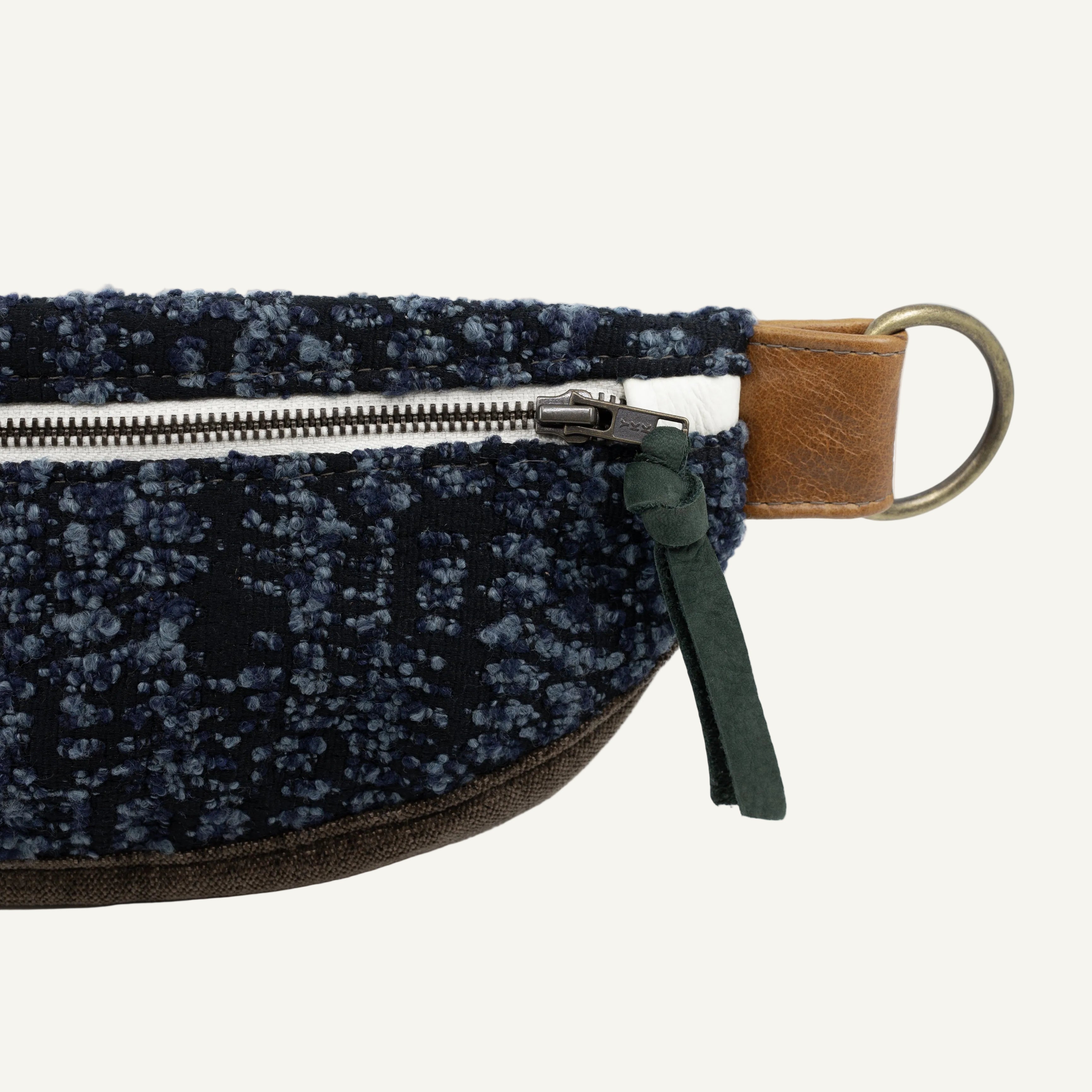 Close-up of a textured dark blue and black speckled fabric pouch with a brown leather zipper pull tab, a brass metal ring, and a green leather zipper pull on a white background.