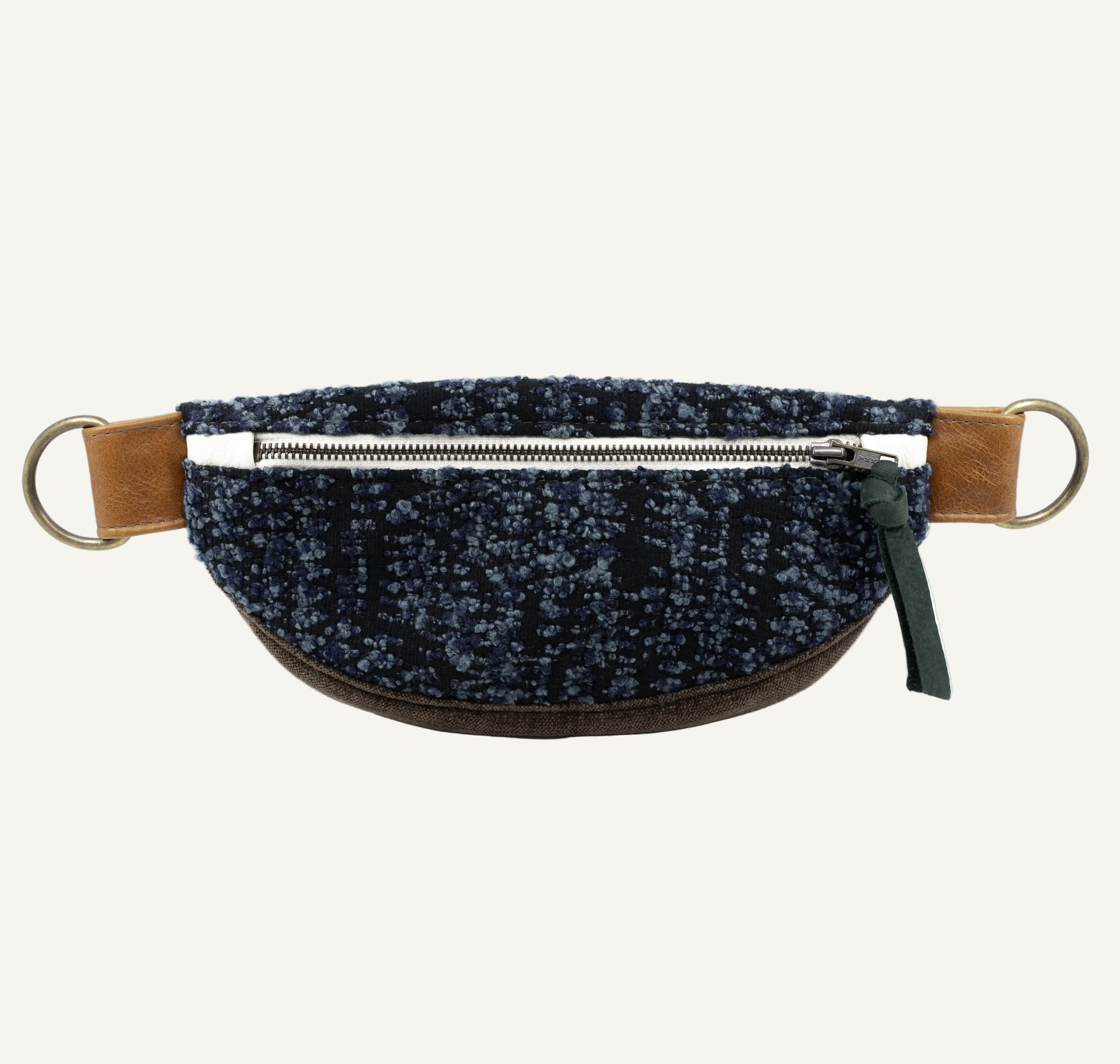 A dark blue and black speckled fabric waist bag, brass hardware, and a green zipper pull, shown against a plain white background.