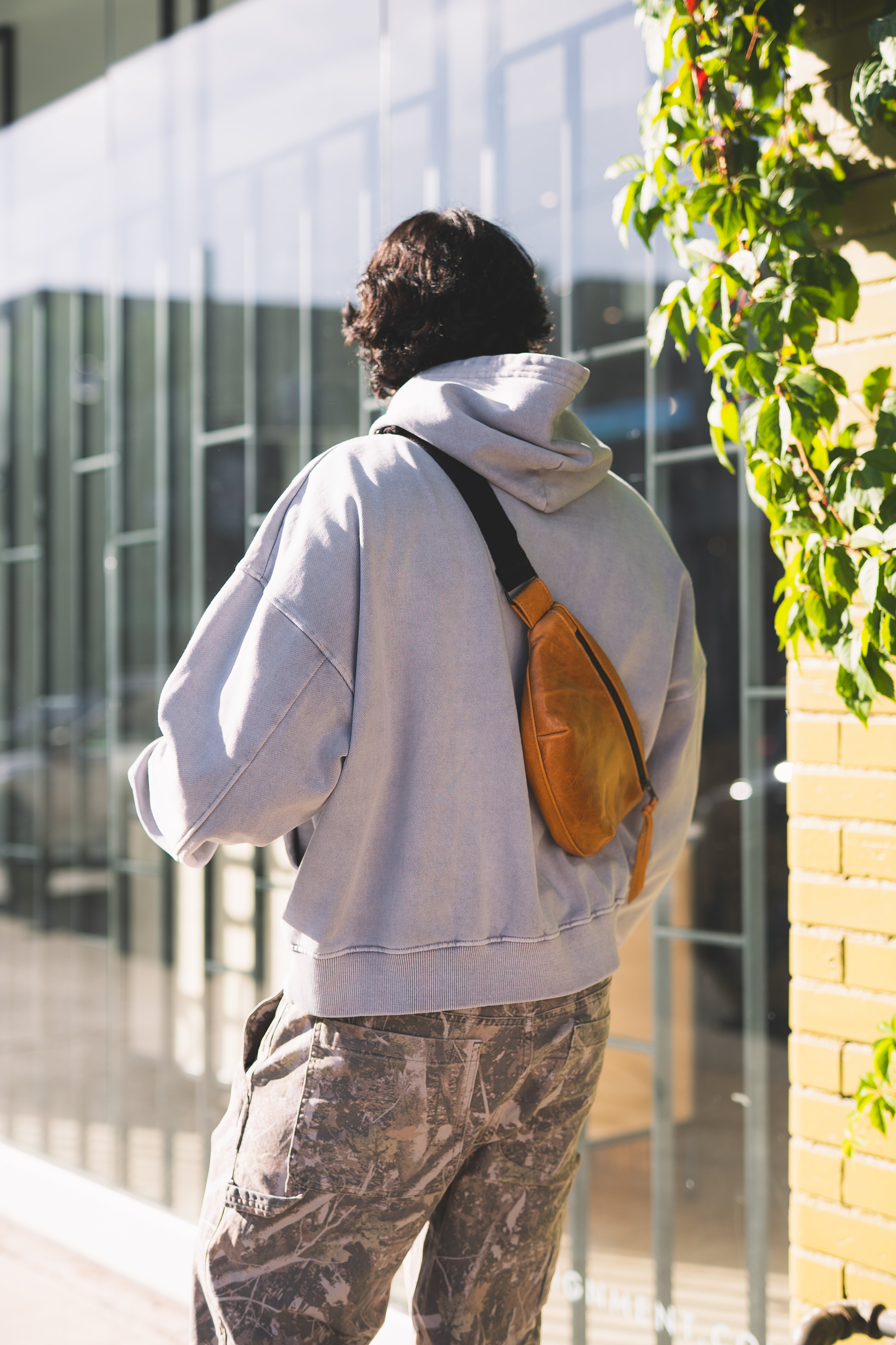 Person wearing a light gray hoodie and camouflage pants with a brown bag, standing outdoors near a building.