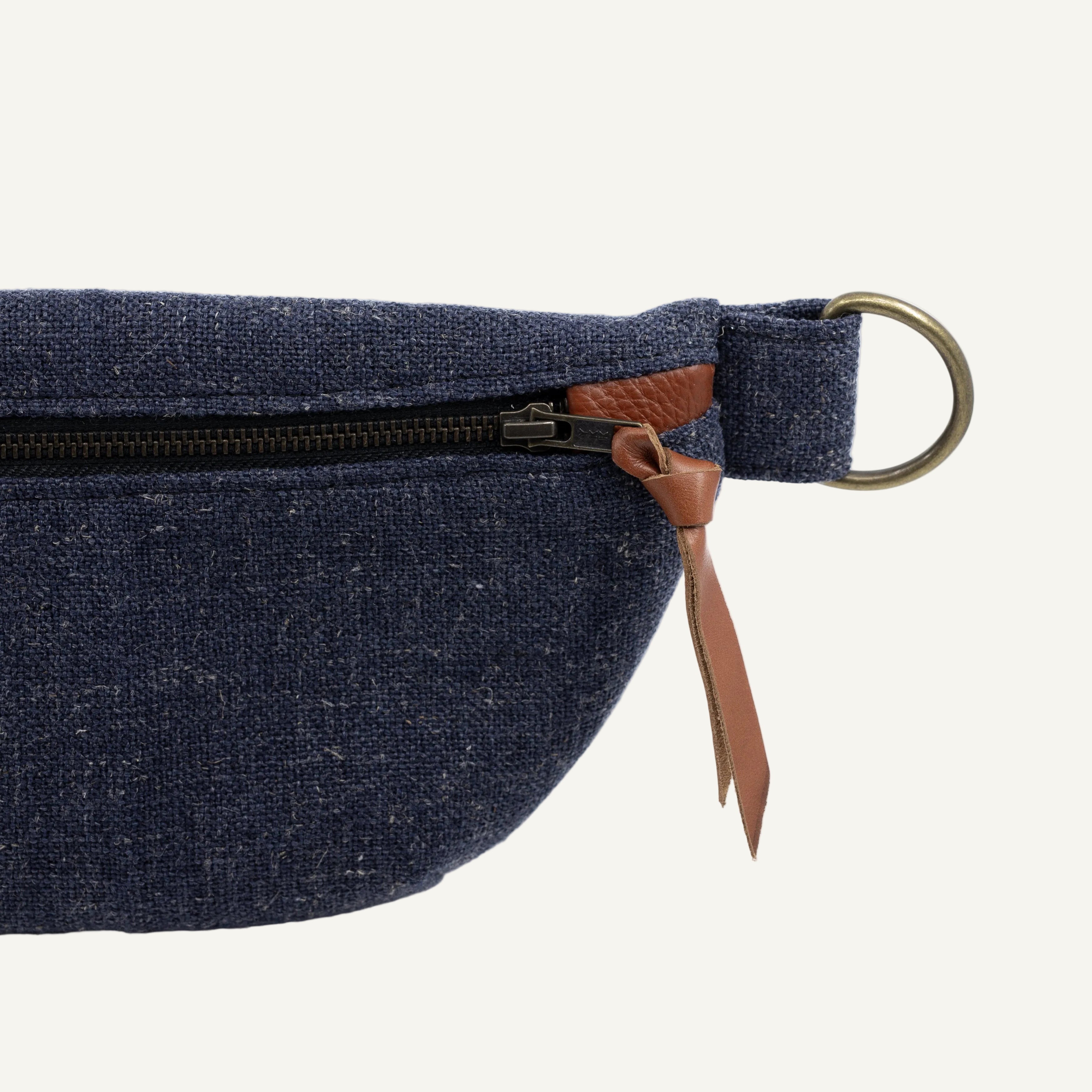 Close-up of a navy blue woven fabric waist bag with a black zipper, brown leather pull tab, and brass ring on a white background.