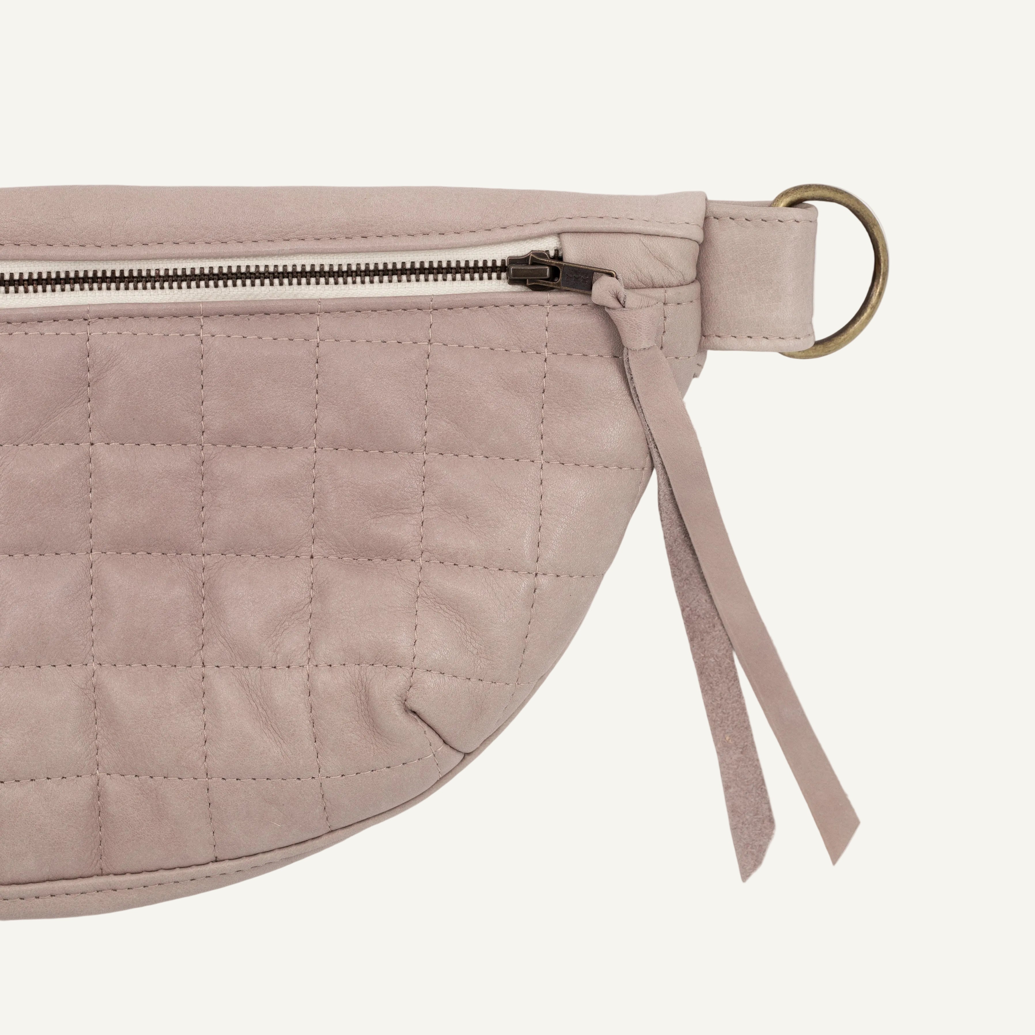 A close-up of a light mauve quilted leather fanny pack with a metal zipper, a brass ring, and a matching zipper pull, shown on a white background.