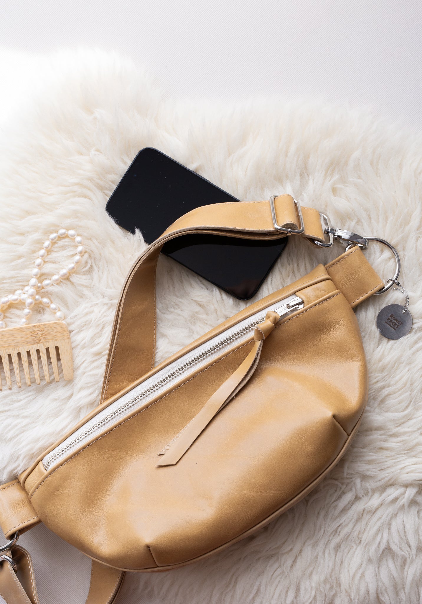 Beige fanny pack on a white fur surface with a phone and hairbrush.
