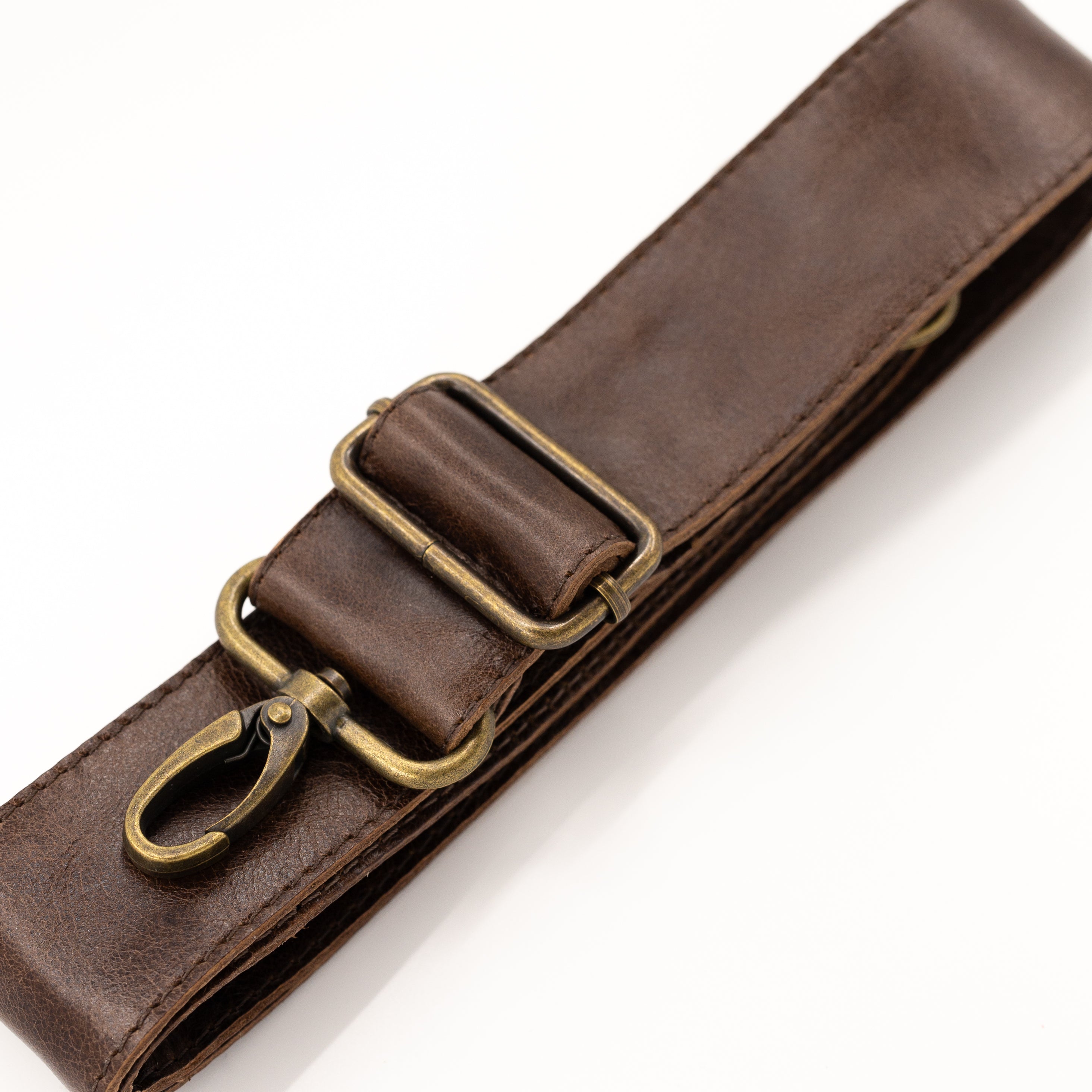 A close-up of a dark brown leather strap with antique brass hardware, including a buckle and a snap hook clasp, set against a plain white background.