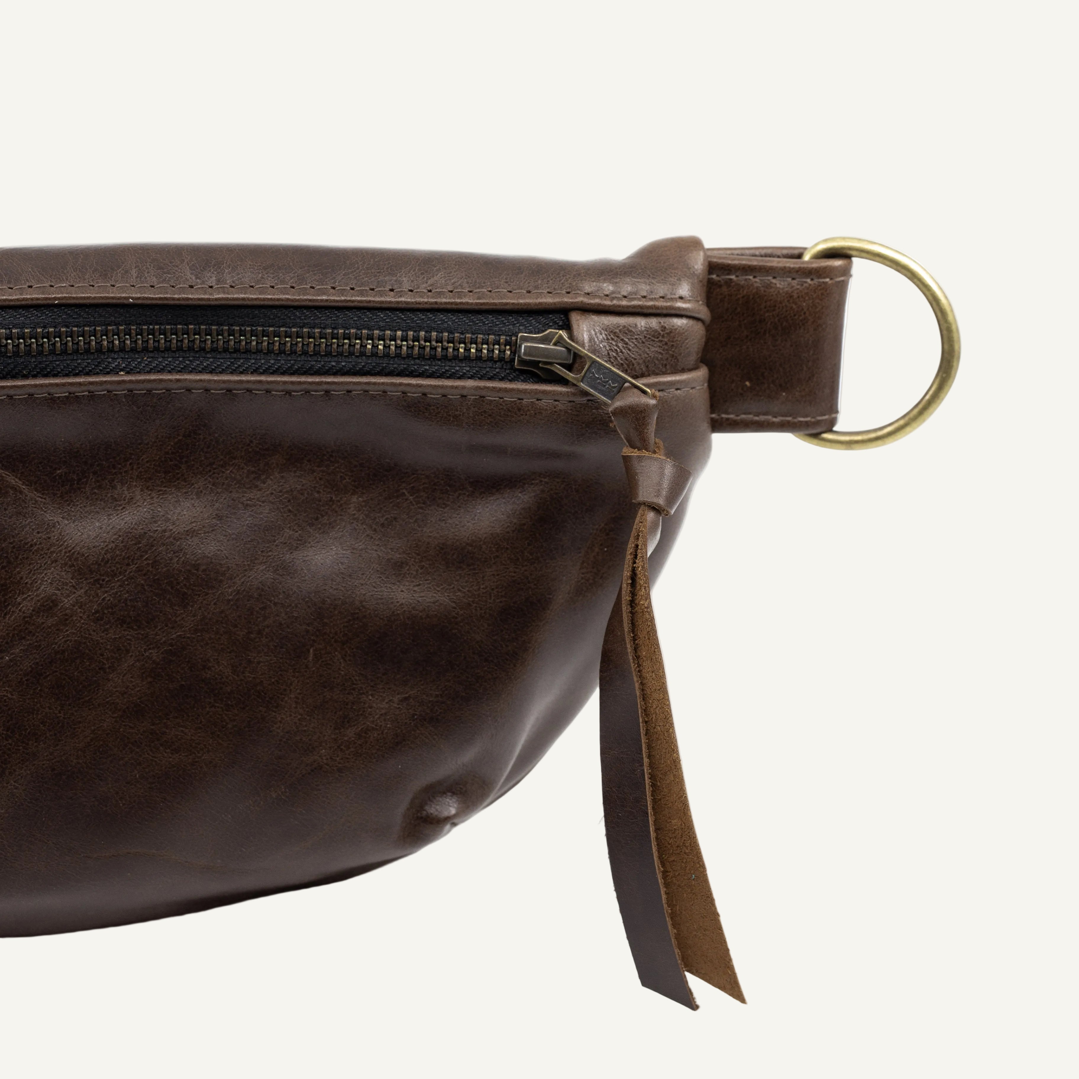 Close-up of a dark brown leather belt bag showing the zipper, and a brass ring on a white background.