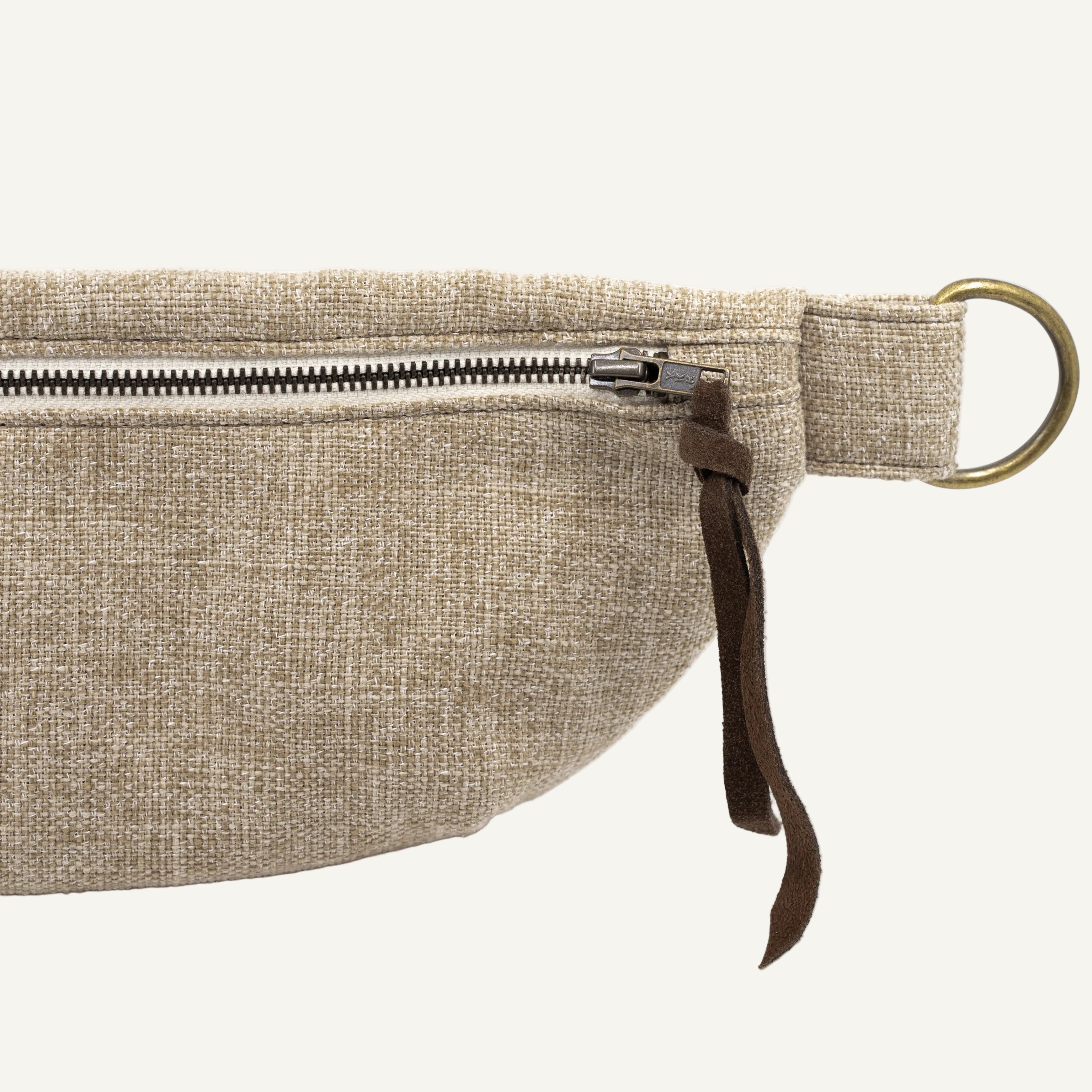 Close-up of a beige woven fabric pouch with a silver zipper, dark brown leather strap tab, and a brass-colored metal ring on a white background.
