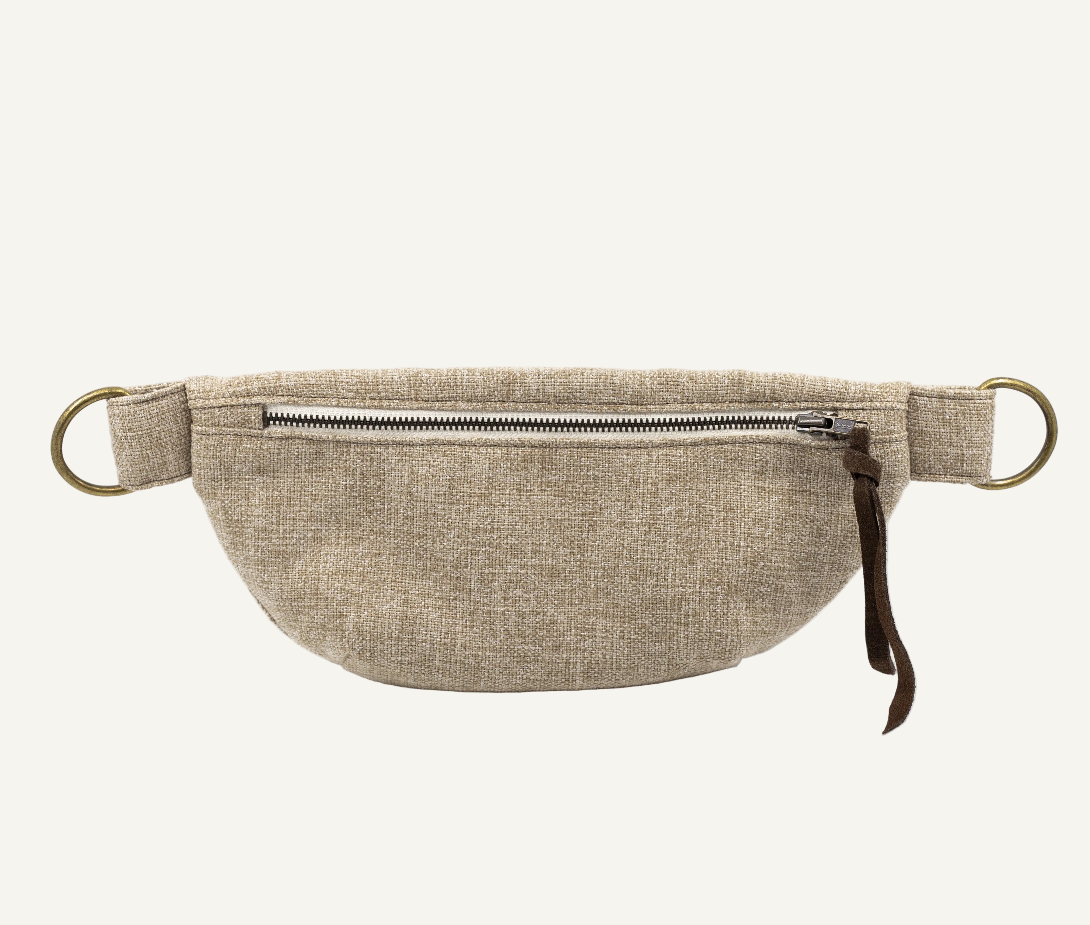 A beige linen waist bag with a front zipper, brass-tone metal rings, and a brown leather pull tab, shown against a plain white background.
