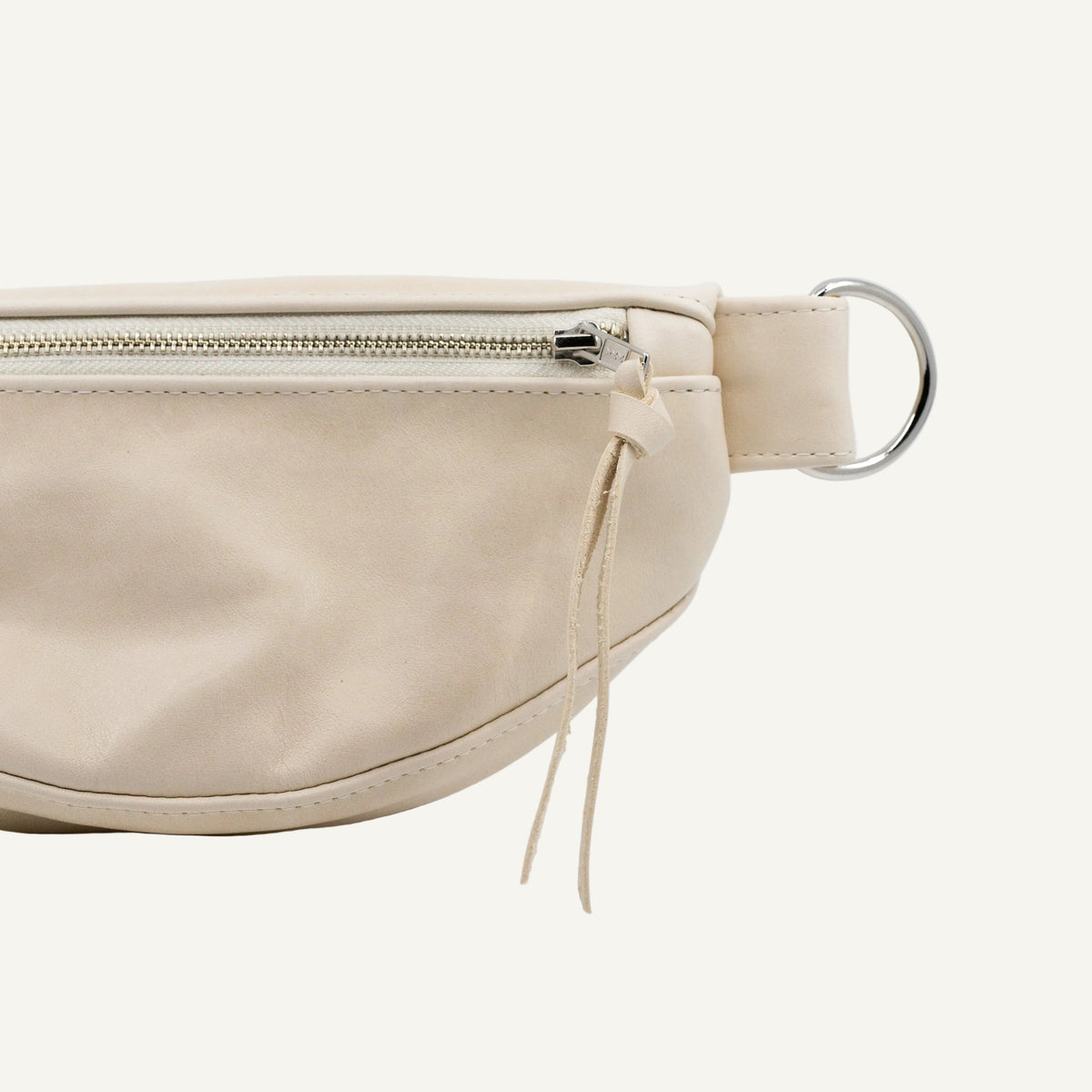 A close-up of a light beige leather belt bag with a silver metal ring attachment and zipper, shown on a clean white background.