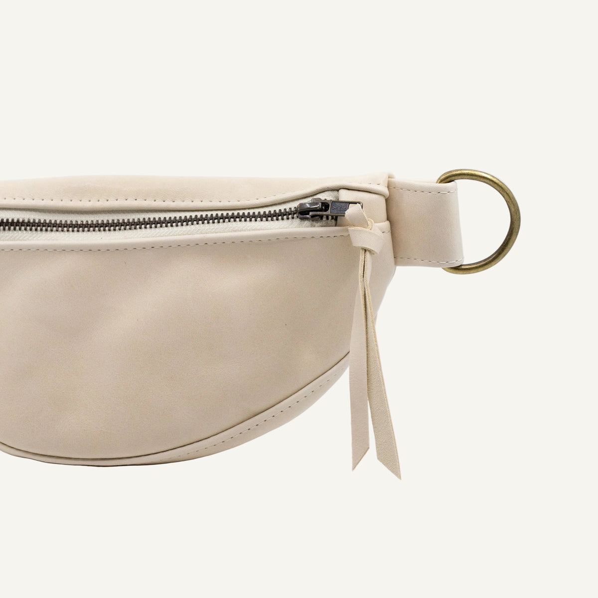 A close-up of a cream-colored leather belt bag with a silver zipper, a gold-tone metal O-ring, and a matching cream leather ribbon tassel, set against a plain white background.