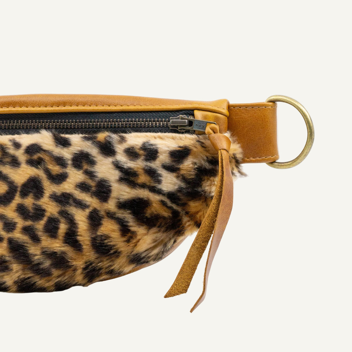 Close-up of a leopard-print faux fur pouch with tan leather trim, a black zipper, and a brass ring on a white background.