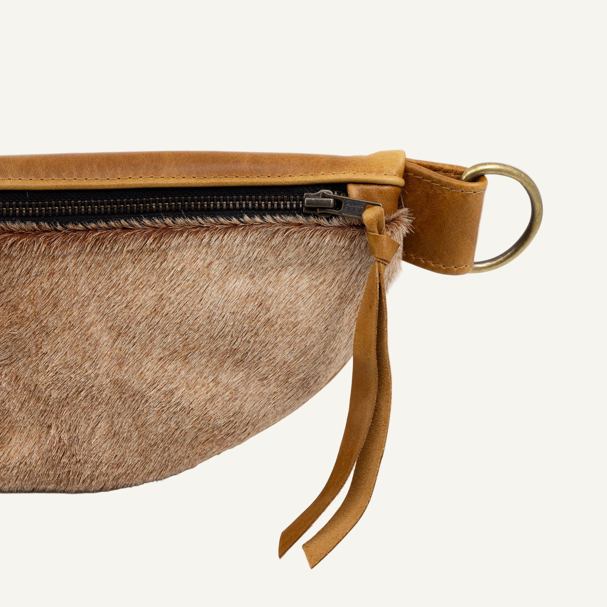 Close-up of a waist bag featuring a tan and brown cowhide body, tan leather trim, a black zipper, and a brass ring on a white background.