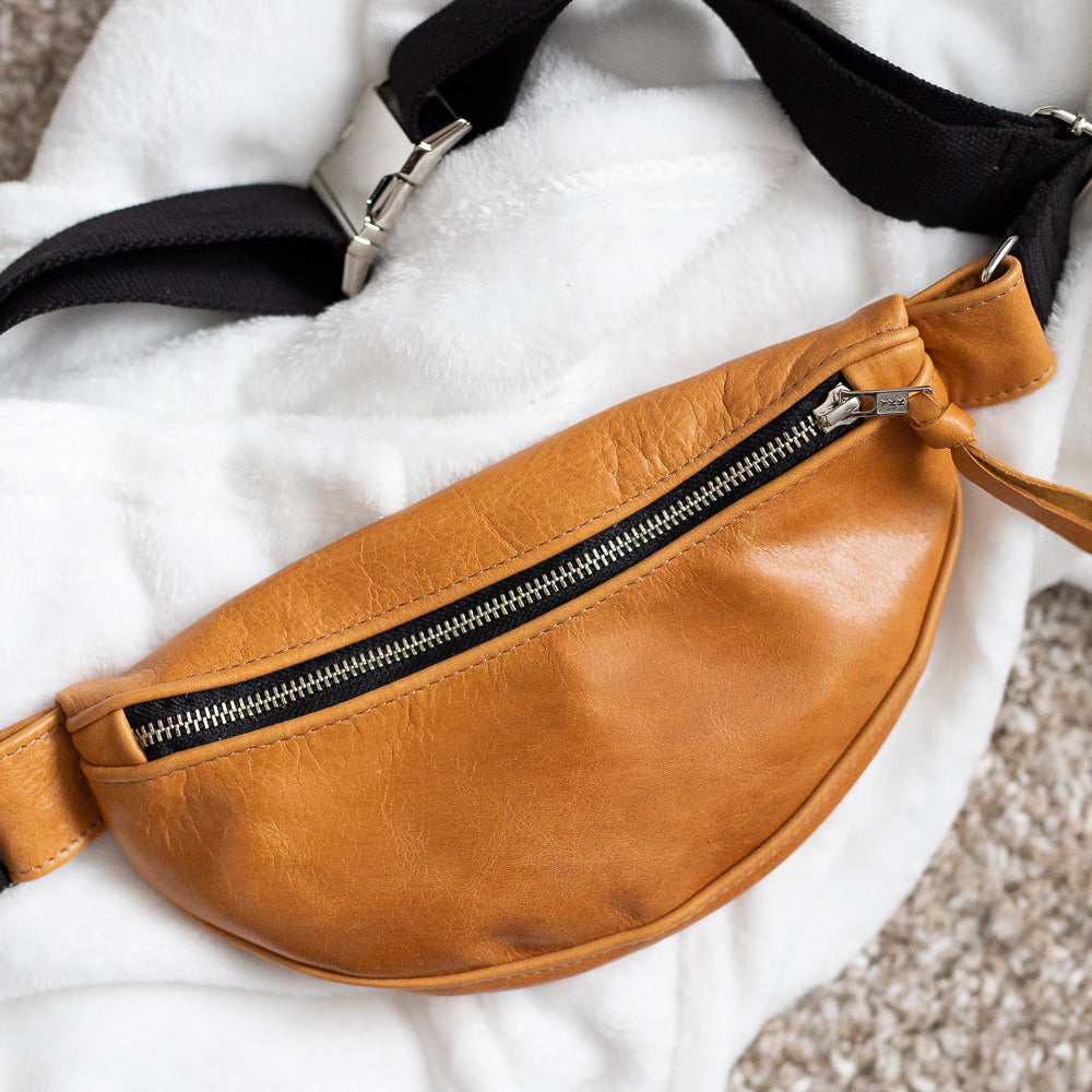 A brown leather fanny pack with an adjustable black strap and metal buckle, displayed on a white fabric surface.
