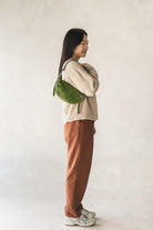Person wearing a beige sweater and brown pants with a green bag on a plain background