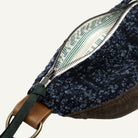 Close-up of a small bag with a dark blue speckled fabric exterior, brown base section, green strap detail, and a cream interior with green printed pattern, shown on a white background.