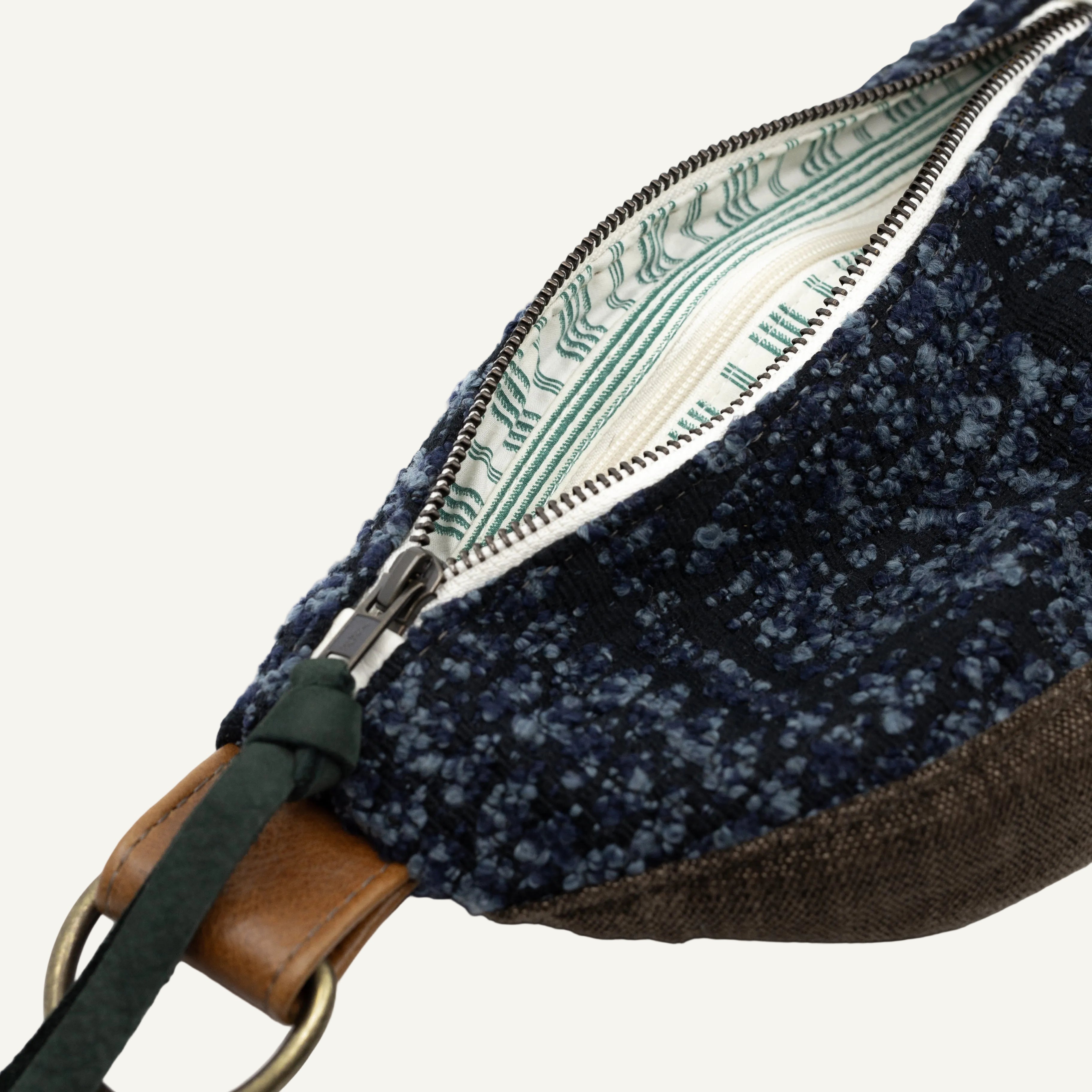 Close-up of a small bag with a dark blue speckled fabric exterior, brown base section, green strap detail, and a cream interior with green printed pattern, shown on a white background.