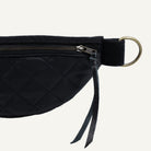 Close-up of a black quilted waist bag with a black strap, brass zipper pull, and a large round metal ring on a white background.
