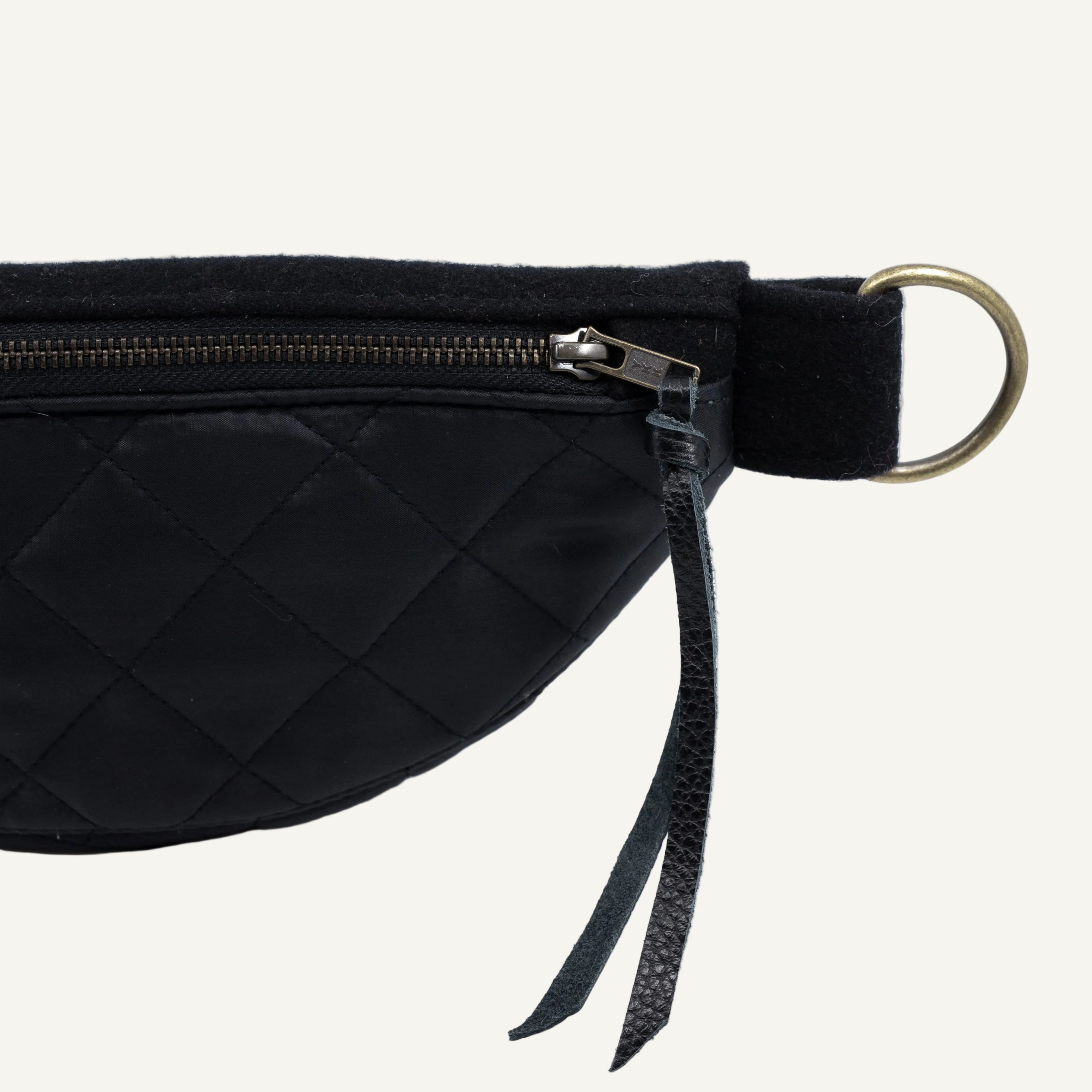 Close-up of a black quilted waist bag with a black strap, brass zipper pull, and a large round metal ring on a white background.