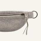 Close-up of a gray textured fabric belt bag with a black zipper, brass-toned metal ring, and a gray fabric pull tab on a white background.