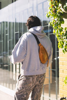 Person wearing a light gray hoodie and camouflage pants with a brown bag, standing outdoors near a building.