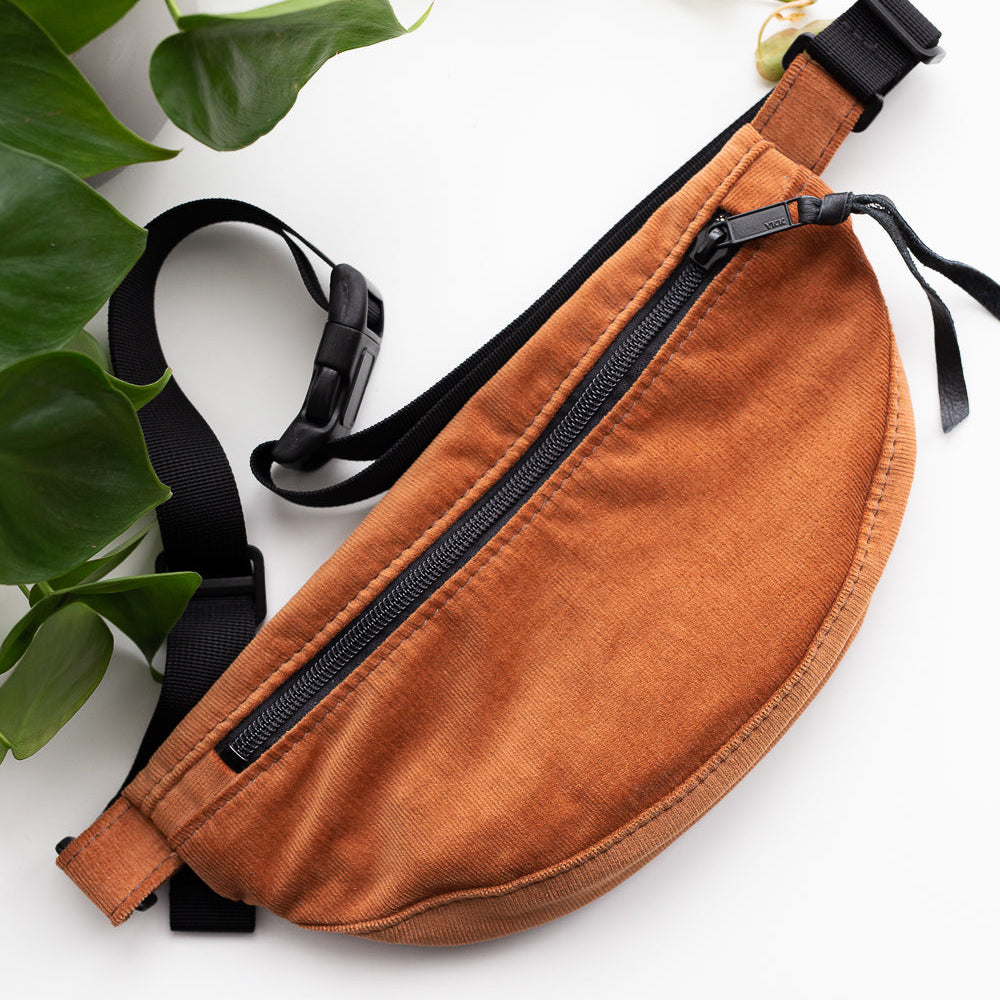 Brown waist bag with black zippers on a white background with green leaves.