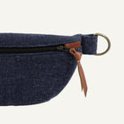 Close-up of a navy blue woven fabric waist bag with a black zipper, brown leather pull tab, and brass ring on a white background.