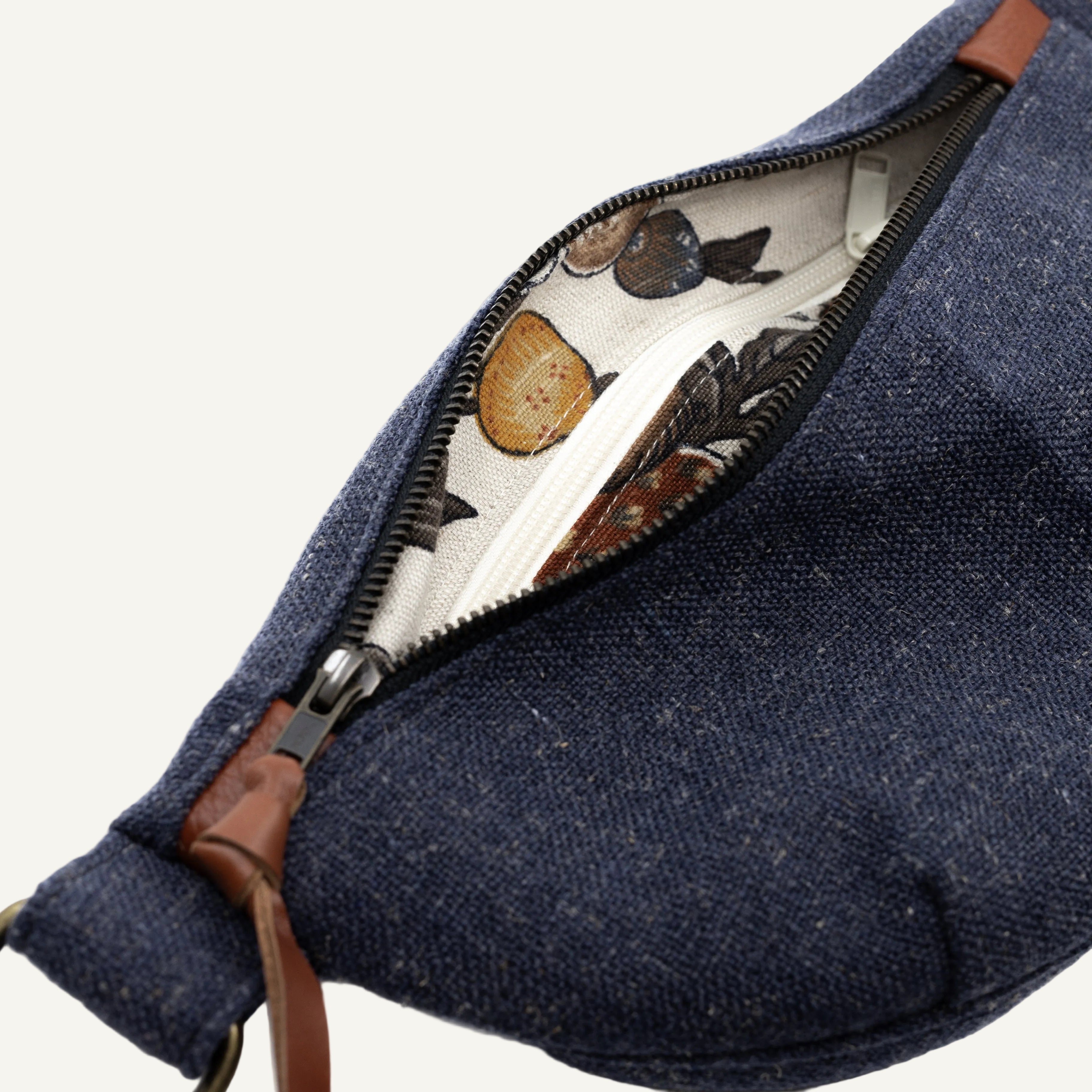 Close-up of a navy blue canvas bag with a partially open zipper, showing a cream-colored interior printed with a vintage-style botanical and bird pattern, and brown leather trim.