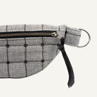 A close-up of a gray and black plaid waist bag or belt bag made of textured fabric, featuring a black zipper, a black leather pull tab, and a silver metal D-ring on a white background.