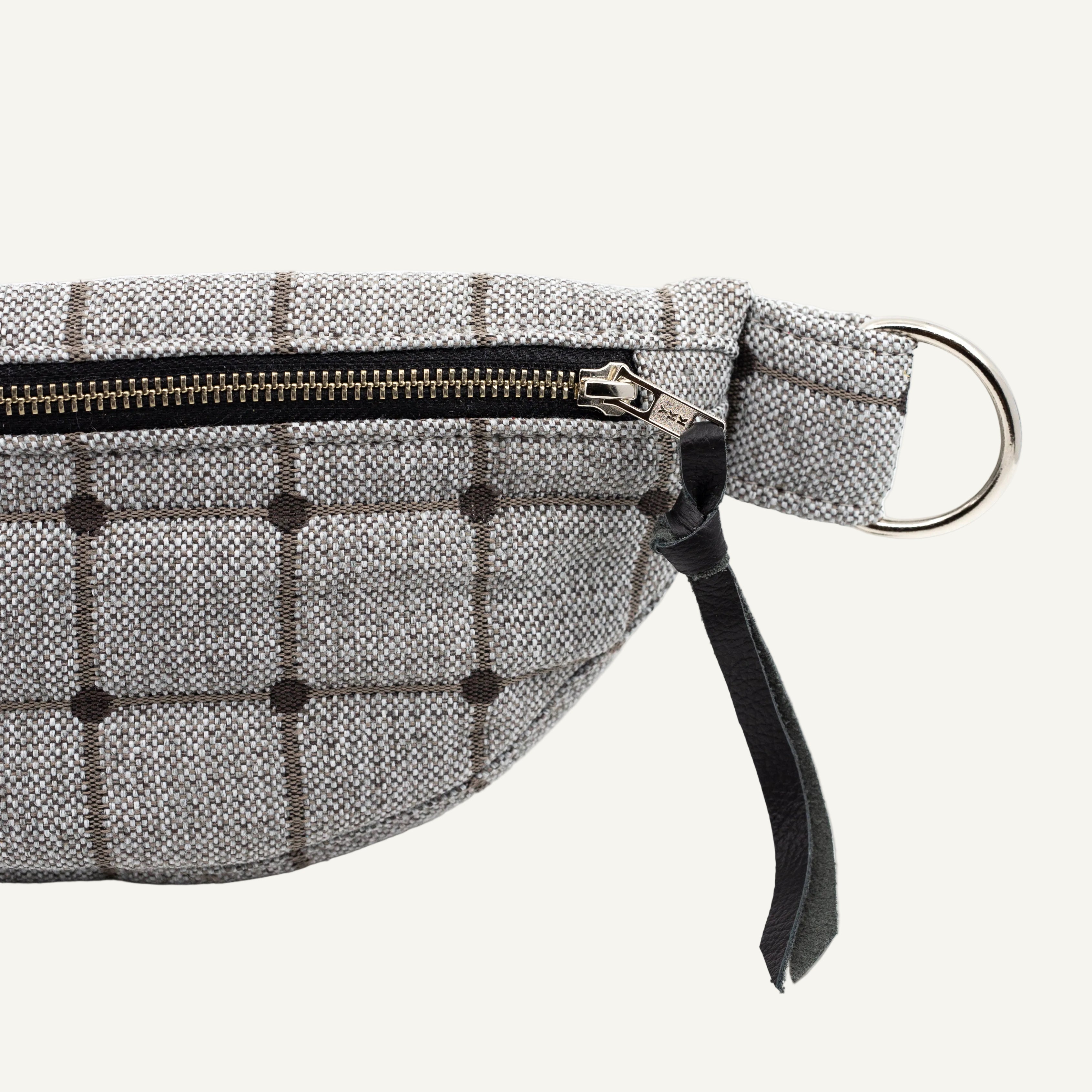 A close-up of a gray and black plaid waist bag or belt bag made of textured fabric, featuring a black zipper, a black leather pull tab, and a silver metal D-ring on a white background.