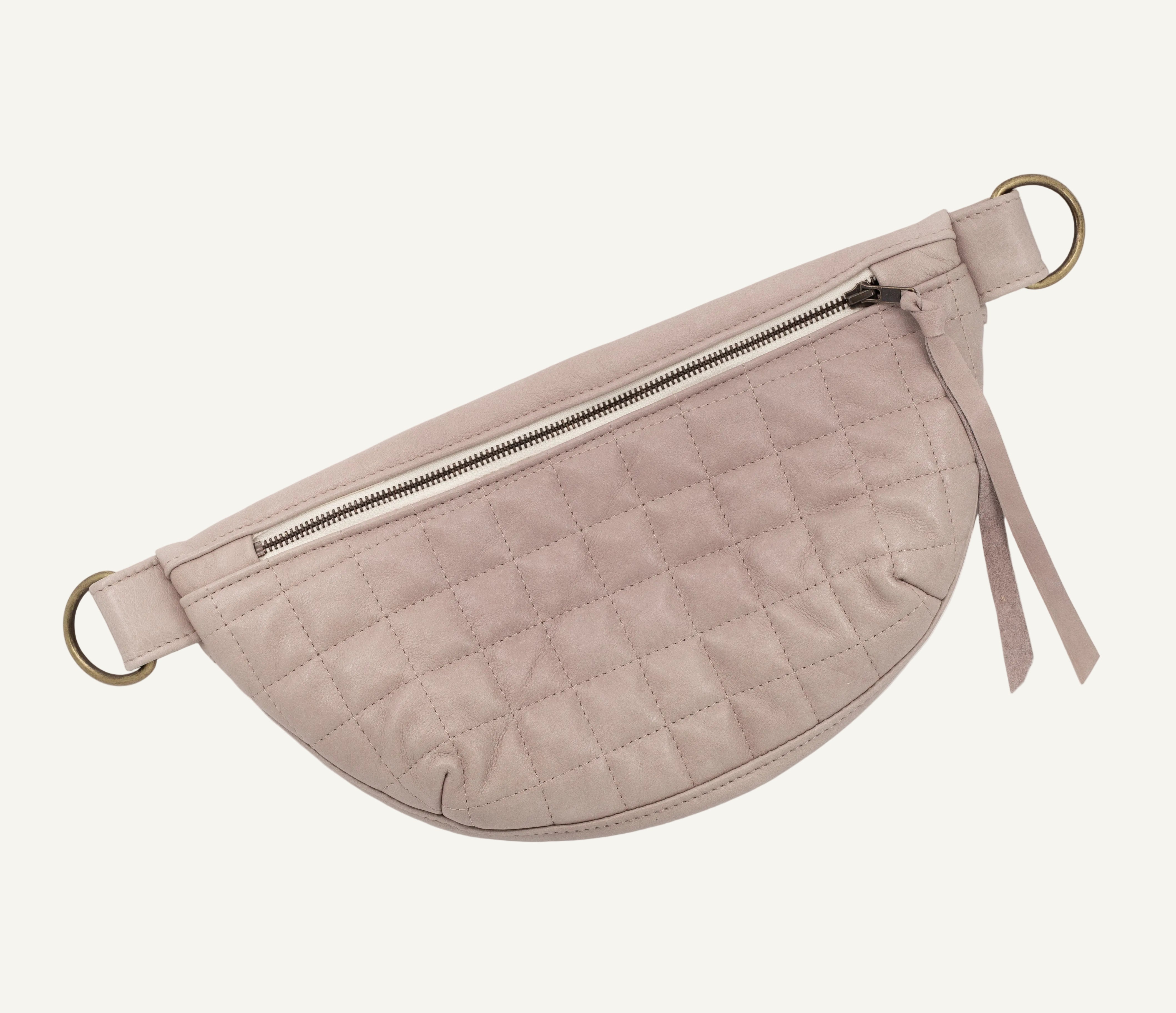 A light mauve quilted leather half-moon fanny pack with a zippered front pocket, and brass-tone hardware, shown on a white background.