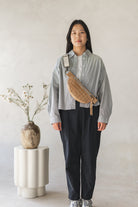 Person wearing a light gray shirt and dark pants with a woven bag, standing against a plain background.