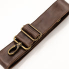 A close-up of a dark brown leather strap with antique brass hardware, including a buckle and a snap hook clasp, set against a plain white background.