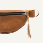 A close-up of a brown leather belt bag showing the side profile, featuring a black zipper, a silver metal D-ring, and a leather pull tab with a gold-tone accent.