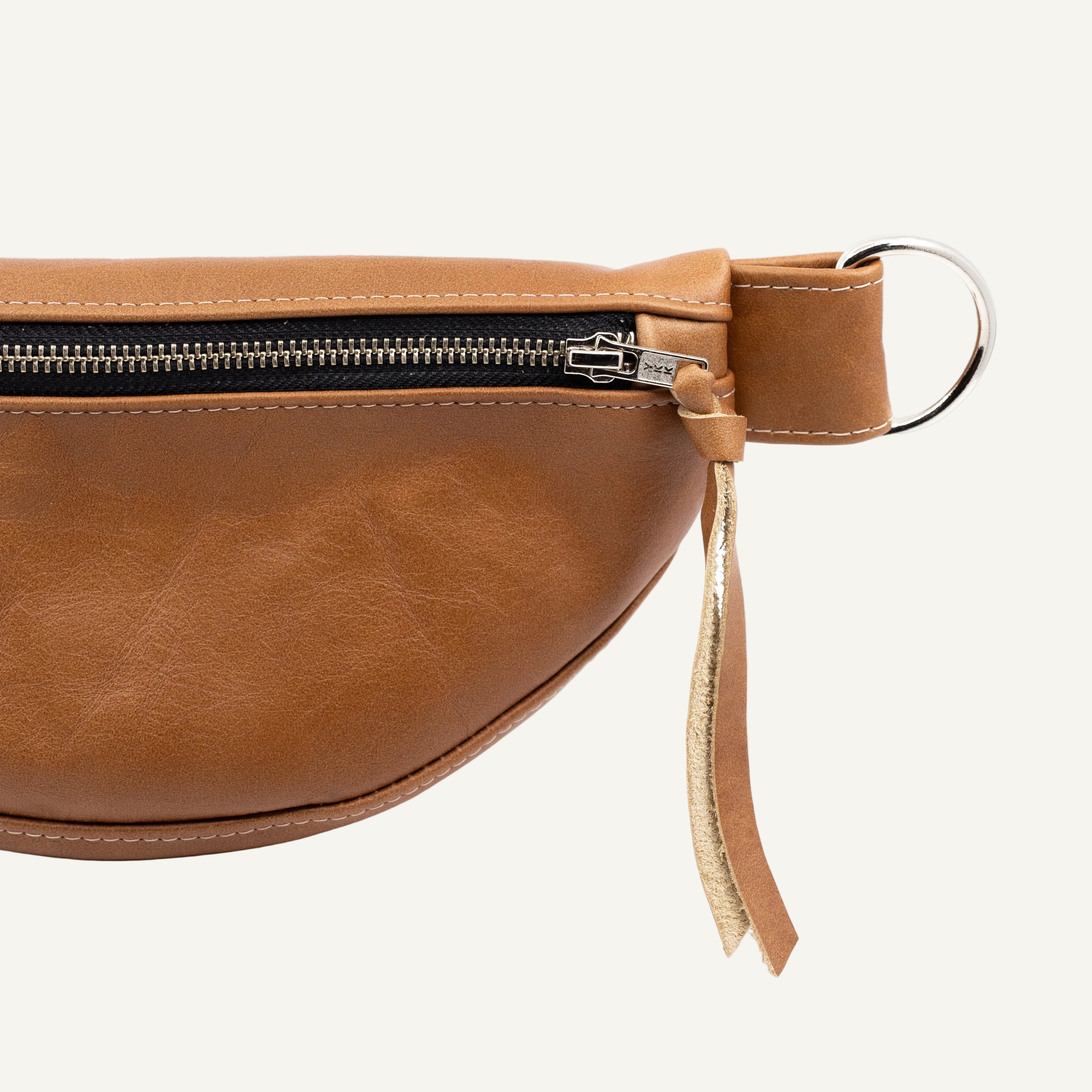 A close-up of a brown leather belt bag showing the side profile, featuring a black zipper, a silver metal D-ring, and a leather pull tab with a gold-tone accent.