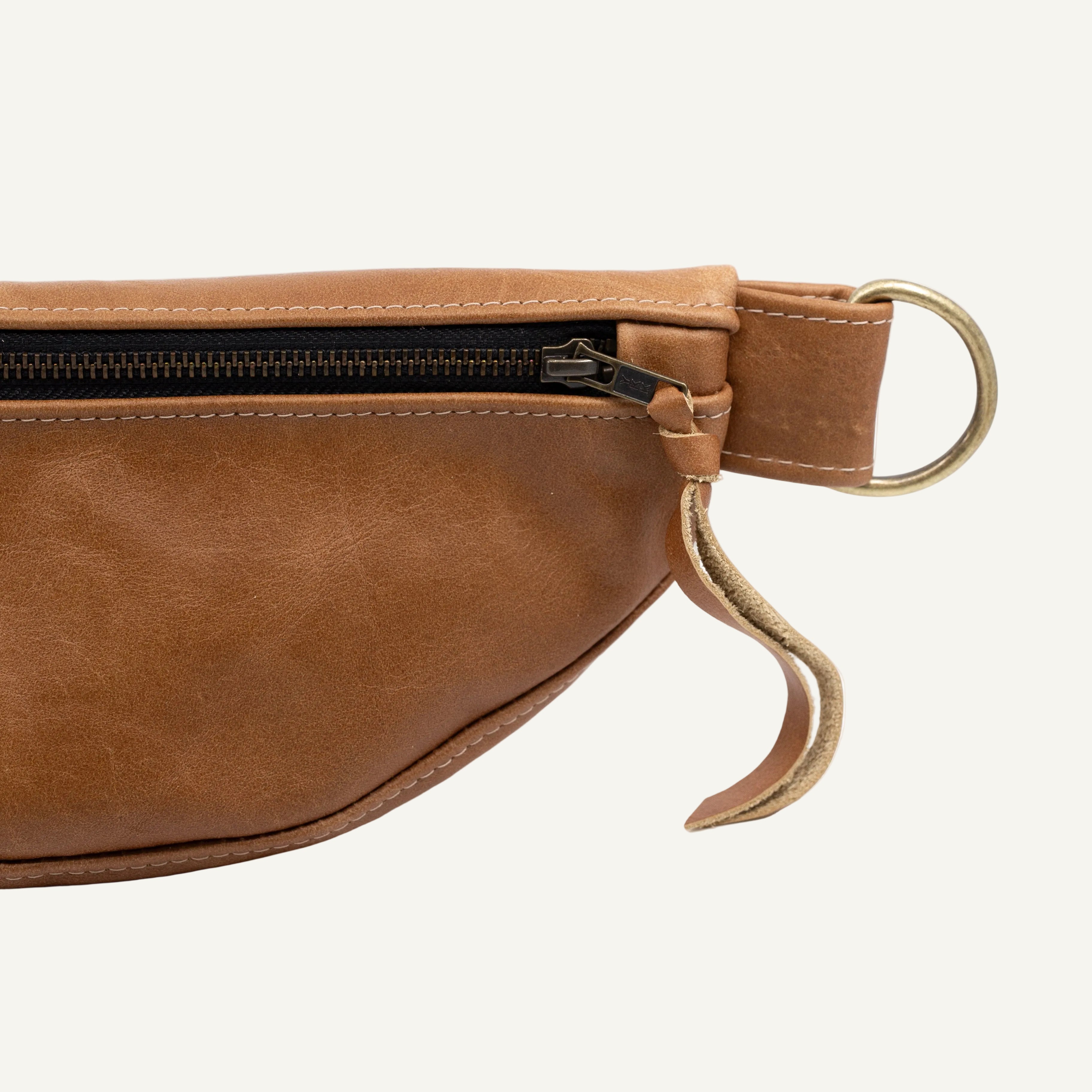 Close-up of a brown leather belt bag showing the side zipper, brass-toned metal ring, detail on a white background.