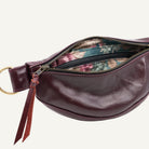 A close-up of a burgundy leather belt bag with a gold-tone ring and zipper, showing a floral-patterned interior lining on a light off-white background.