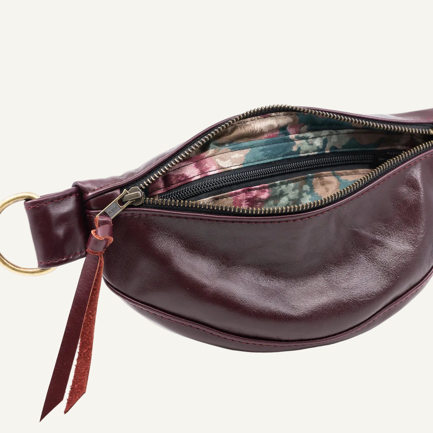 A close-up of a burgundy leather belt bag with a gold-tone ring and zipper, showing a floral-patterned interior lining on a light off-white background.