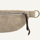 Close-up of a beige woven fabric pouch with a silver zipper, dark brown leather strap tab, and a brass-colored metal ring on a white background.