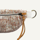 A close-up of a textured tweed waist bag in a beige, white, and rust-brown pattern, featuring a brass hardware.