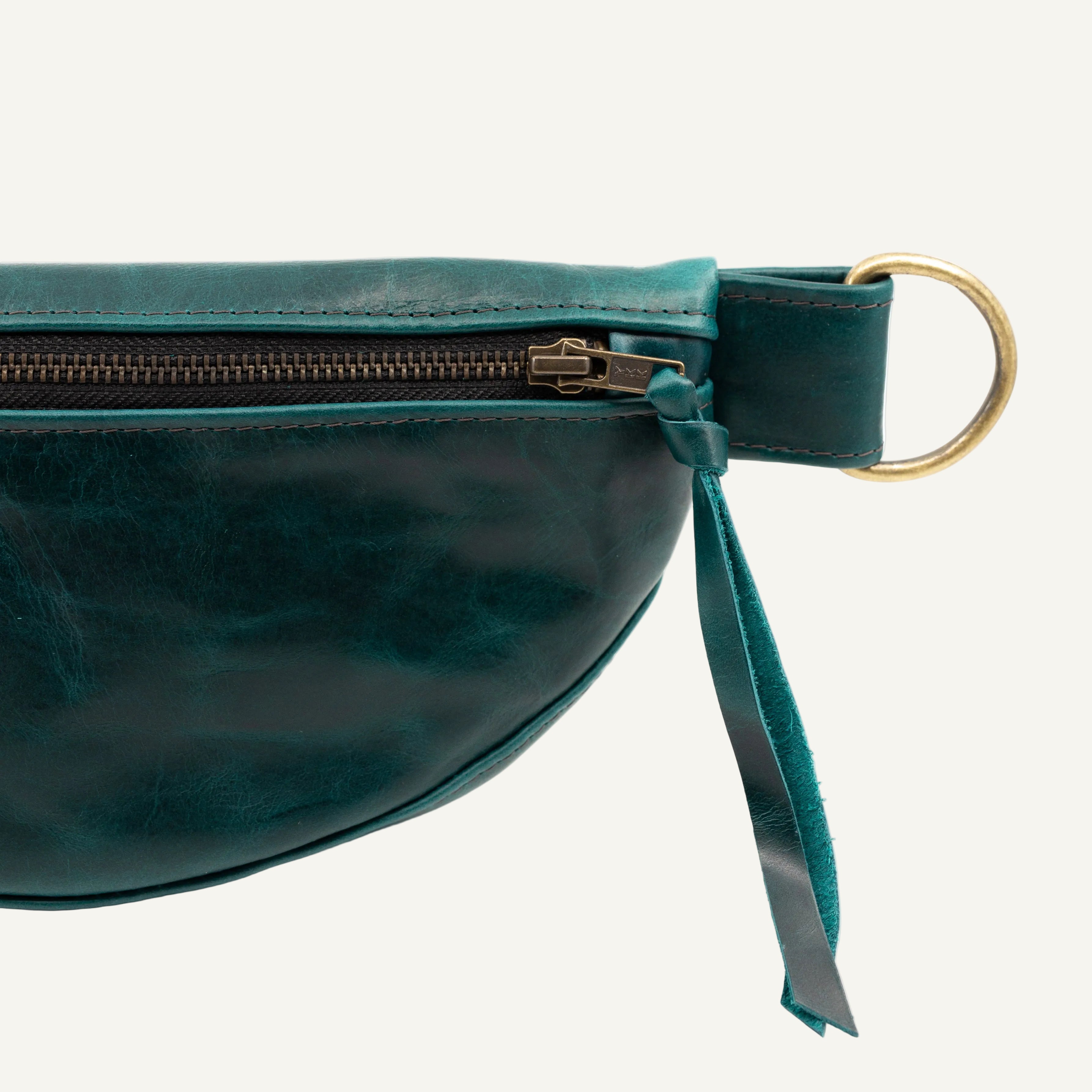 Close-up of a dark teal leather belt bag showing a curved front, a dark zipper with metal hardware, on a white background.