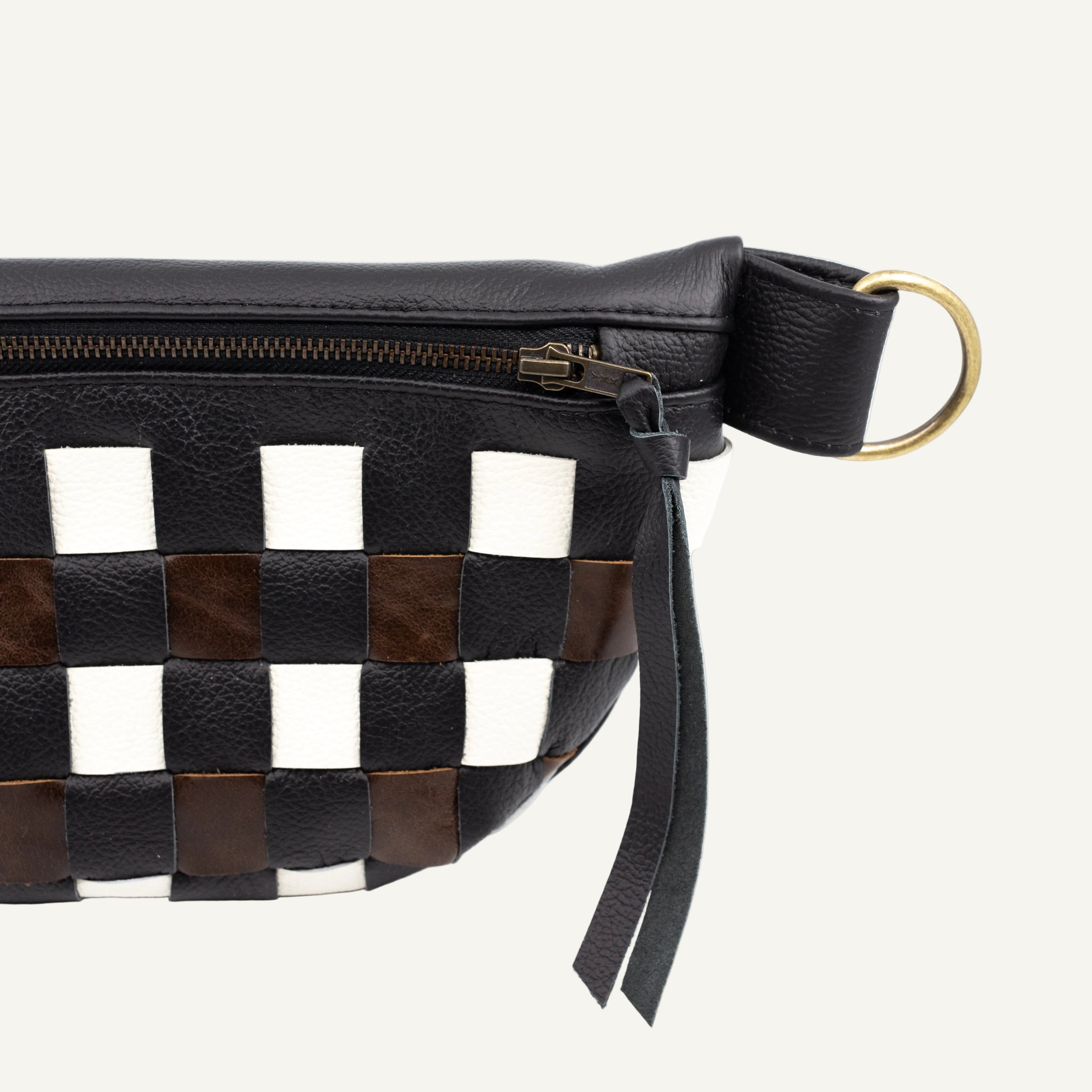 Close-up of a black leather belt bag with a woven checkerboard pattern in black, white, and brown, featuring a gold-tone ring clasp and a black leather zipper pull.