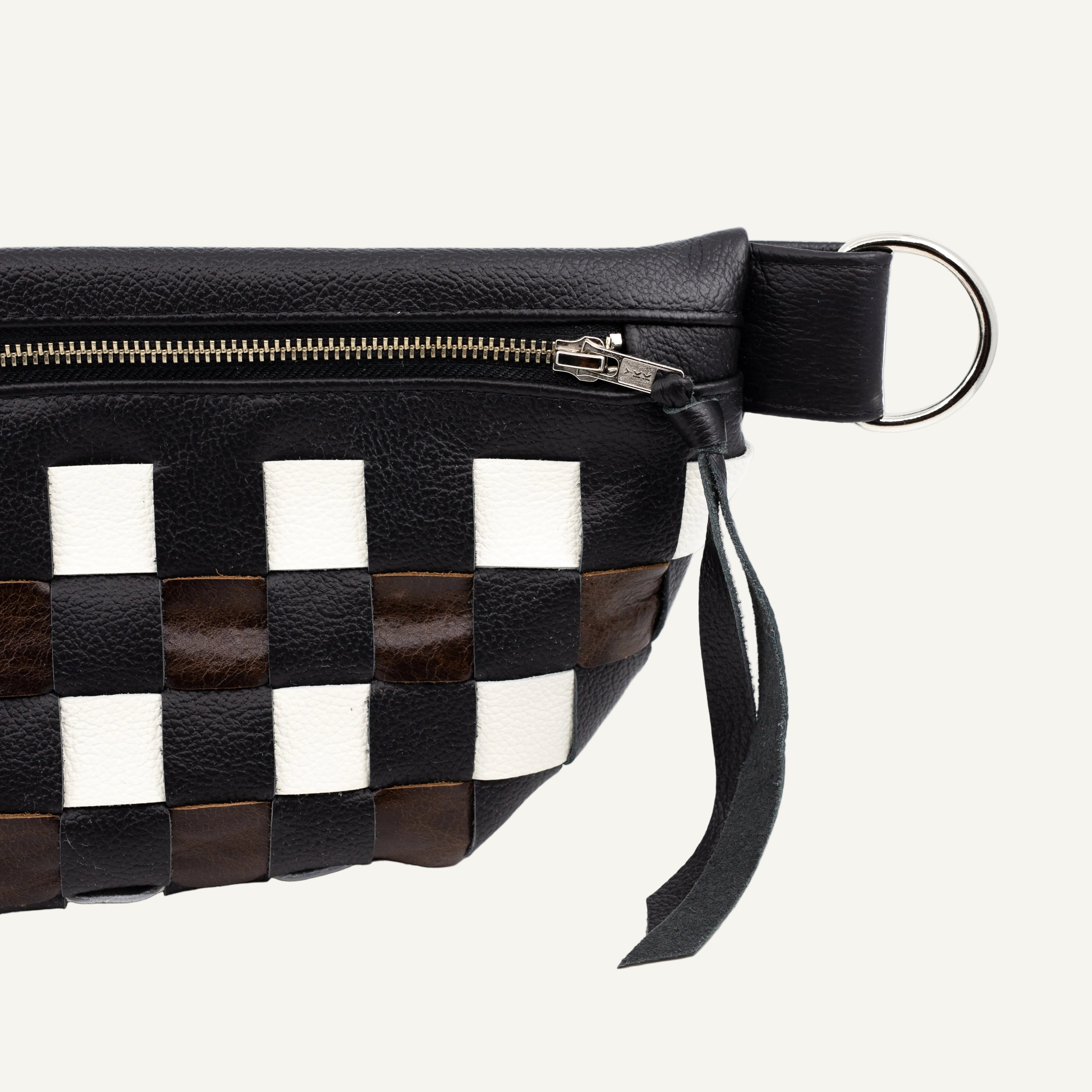 A close-up of a black leather pouch featuring a woven checkerboard pattern with black, white, and dark brown leather strips, a chrome zipper, with a silver metal ring.