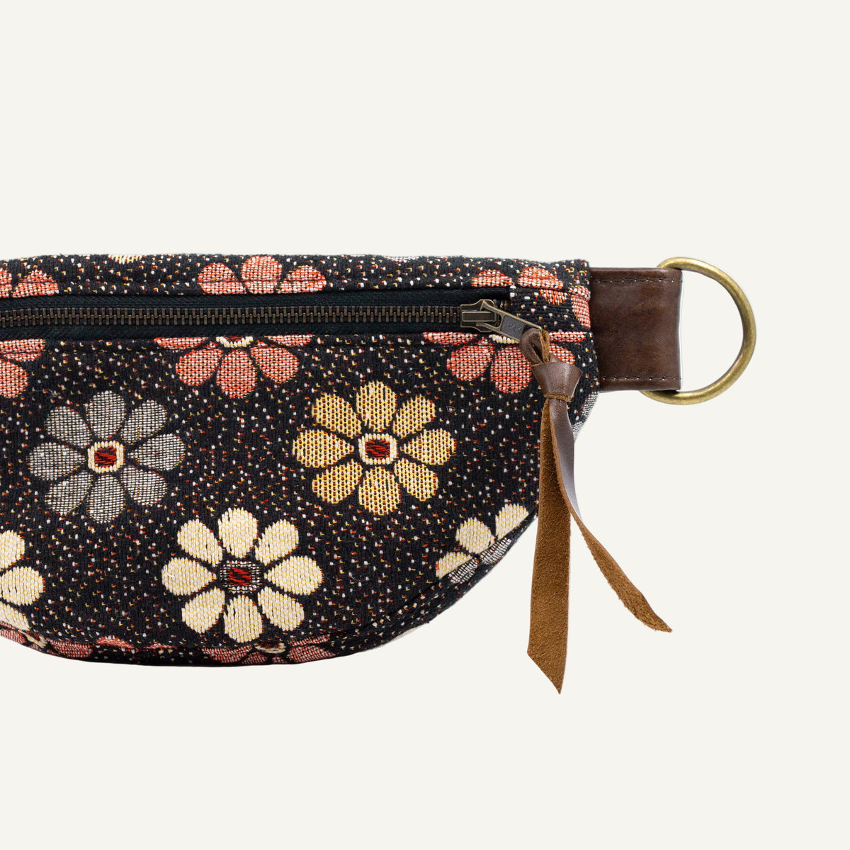 A close-up of a waist bag featuring a black fabric body with a colorful embroidered floral pattern in pink, beige, and gray. It has a brown leather strap, a brass ring, and a brown leather tassel zipper pull.