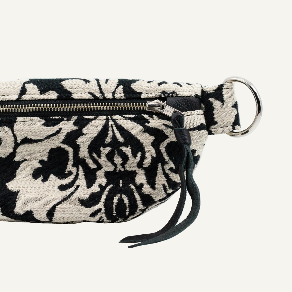 A close-up of a black and cream damask patterned fabric pouch with a gold zipper, a dark green leather tassel, and a silver metal O-ring on a white background.