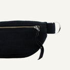 Close-up of a black ribbed fabric waist bag with a silver zipper, black strap, and silver metal ring on a light off-white background.