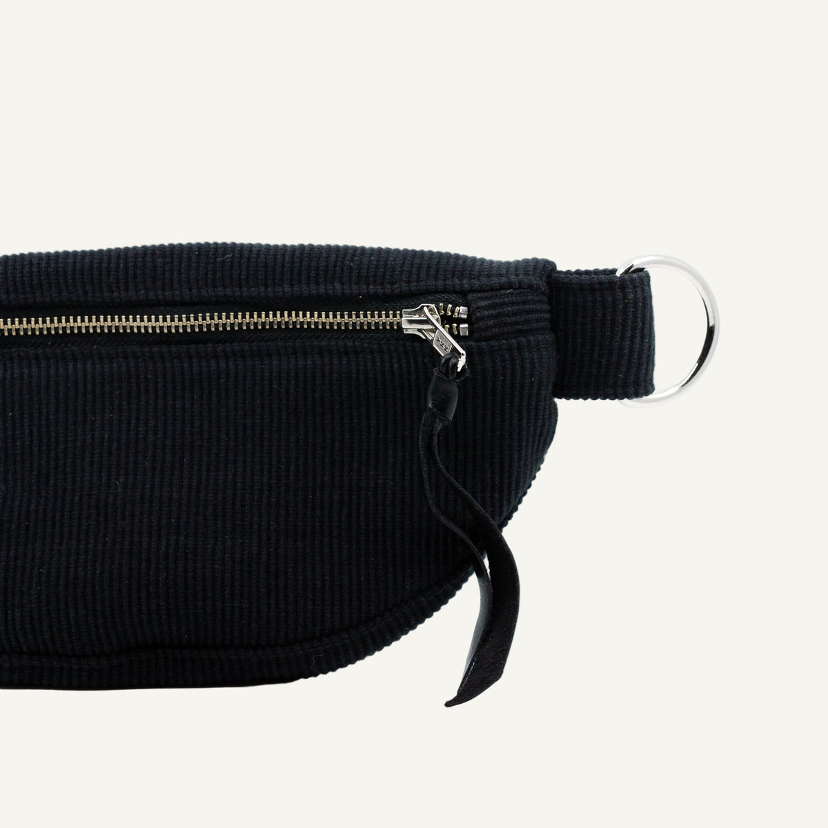 Close-up of a black ribbed fabric waist bag with a silver zipper, black strap, and silver metal ring on a light off-white background.