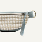 Close-up of a small belt bag with a woven beige and cream body, a muted blue-gray leather trim and strap, and a brass ring hardware on a white background.