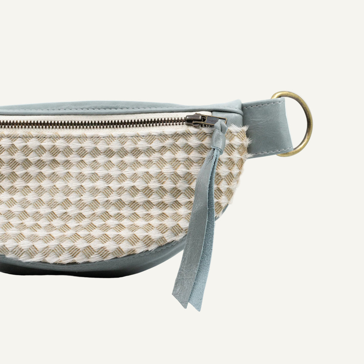 Close-up of a small belt bag with a woven beige and cream body, a muted blue-gray leather trim and strap, and a brass ring hardware on a white background.