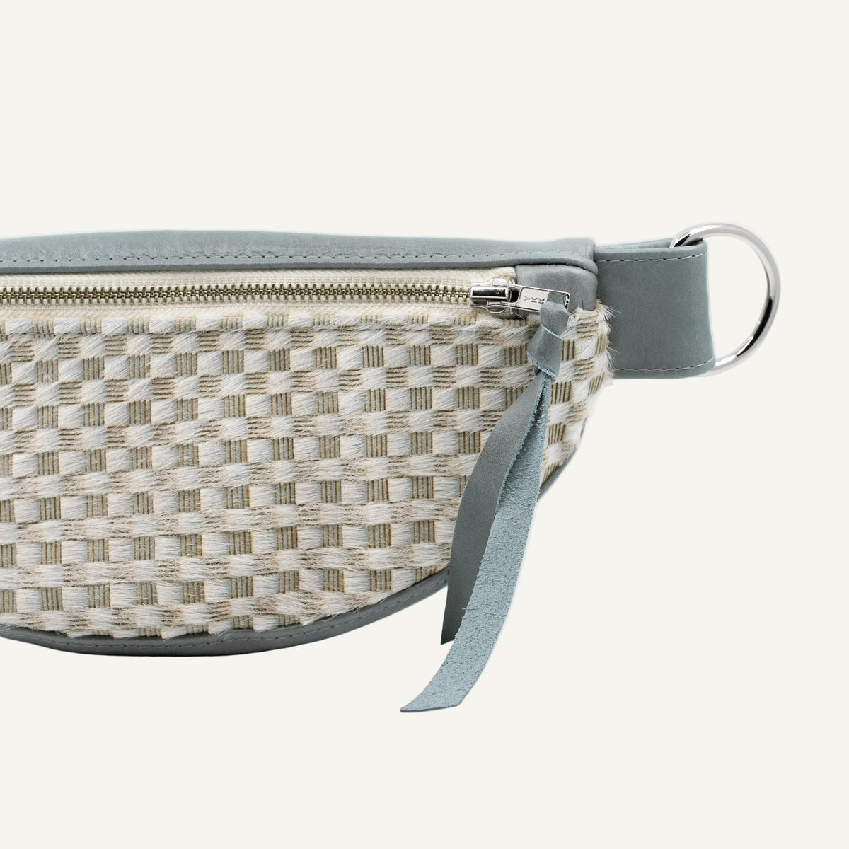 A close-up of a belt bag featuring a cream and beige pattern, a muted blue-gray leather strap and zipper, and a silver metal ring.