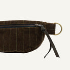 A close-up of a dark brown pinstriped fabric belt bag with a black zipper, black leather strap, and a brass ring on a white background.