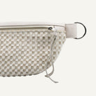 A close-up of a belt bag in beige and off-white, featuring a silver zipper, leather strap, and a silver metal O-ring on a white background.