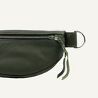 An olive green leather belt bag shown in a close-up side view, featuring a silver metal zipper, a silver D-ring, and a matching leather pull tab on a white background.